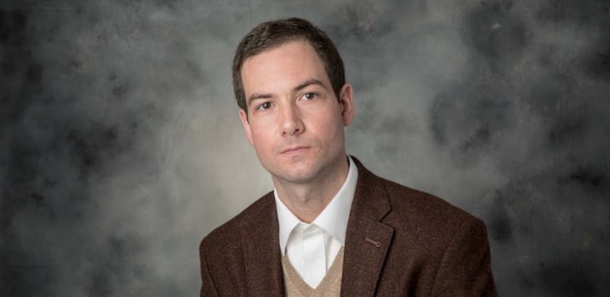 Adrian Baez-Ortega, in a brown blazer, against a blackboard background