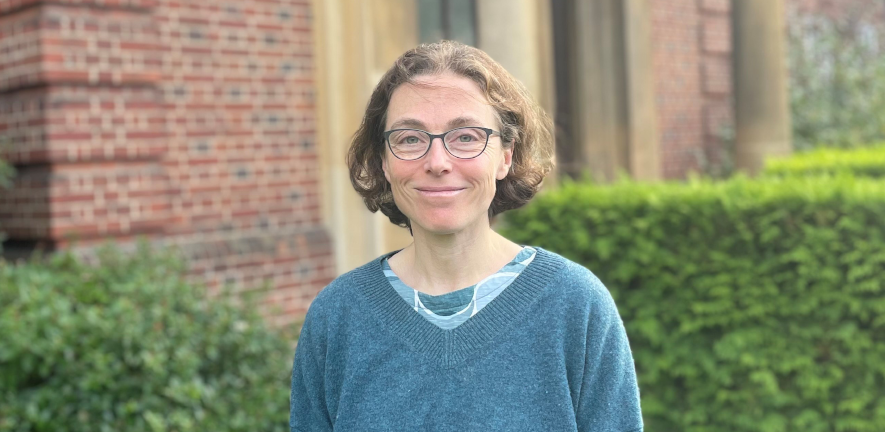 Kate D Baker, smiling, with short brown hair and glasses, in front of blurred background of a building and garden