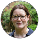 Kate S. Baker, smiling, with hair up, wearing glasses, a blue shirt and DNA earrings, in front of blurred garden background