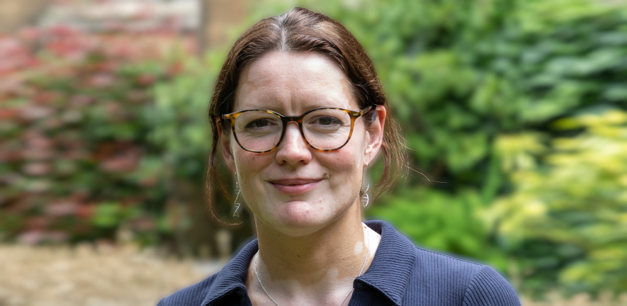 Kate S. Baker, smiling, with hair up, wearing glasses, a blue shirt and DNA earrings, in front of blurred garden background