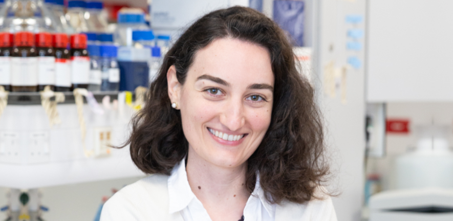 Woman with curly brown hair smiling, stood in a laboratory 