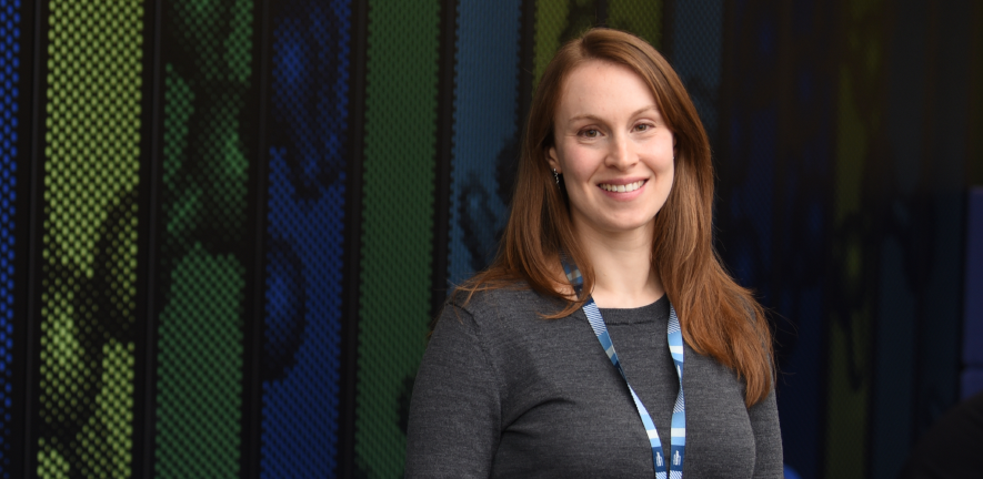 Erica Bello, smiling, in grey top with blue lanyard, in front of green and blue striped background