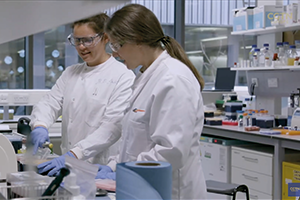 scientists wearing lab coats, goggles and gloves looking at equipment on lab bench