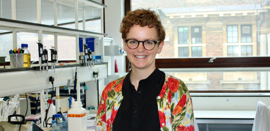 Camilla Godlee, smiling, standing in the laboratory wearing a floral cardigan, glasses, and with short hair