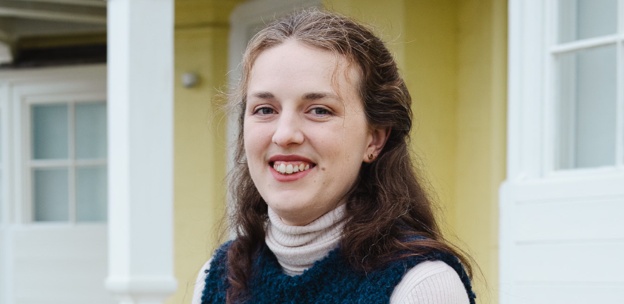 Iris Hardege, smiling, in a blue jumper, in front of a yellow building