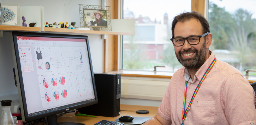 Adrian Liston, smiling, sat at his desk next to a computer displaying data