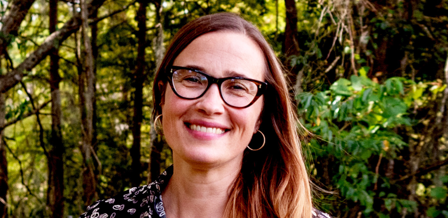 Christine Miller, smiling, in a black shirt and glasses against a forest backgroudn