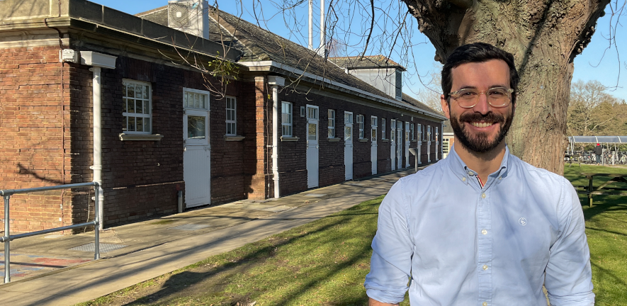 Jose Novo Matos, in a shirt, stood smiling in front of a tree and a brick building 