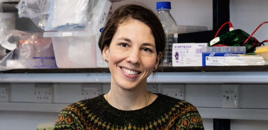 Jeanne Salje, smiling, in a knitted jumper, standing in her lab 