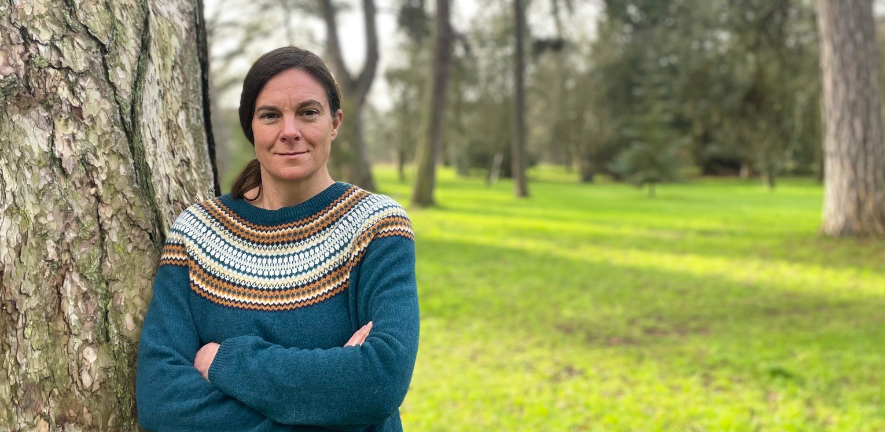 Katharina Schiessl, smiling, arms crossed, wearing blue jumper, against a tree in the botanic gardens 