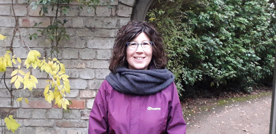 Debra Spencer wearing glasses and a purple outdoor jacket, standing in front of an arch and greenery