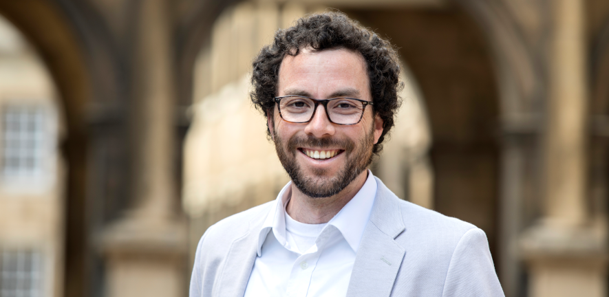Head and shoulders shot of Andrew Tanentzap, wearing glasses, a white shirt and a blue suit jacket in front of a blurred background of an old archway 