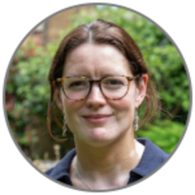 Kate S. Baker, smiling, with hair up, wearing glasses, a blue shirt and DNA earrings, in front of blurred garden background